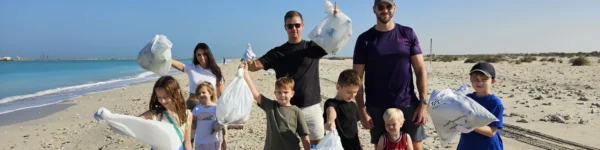 A group of adults and children stands on a sandy beach, holding bags of collected litter, with a clear blue sea in the background.