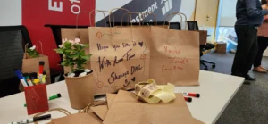 Table with decorated paper bags, potted plants, and markers, showcasing personalized notes and a warm, collaborative atmosphere.