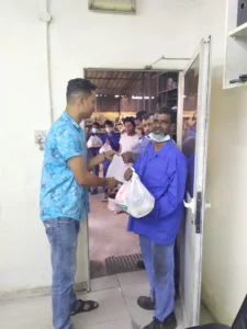 A smiling man in blue receives a bag from another man at a door, with a group of people visible in the background.