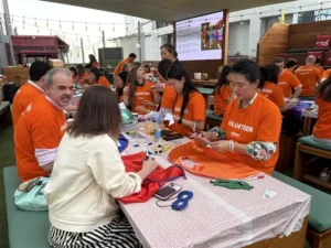 A group of volunteers in bright orange shirts engages in crafting activities at a communal table, surrounded by supplies and decorations.
