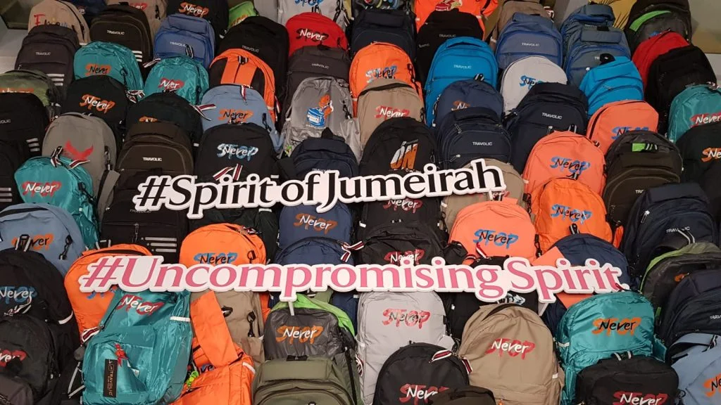 Several colorful backpacks are displayed in rows with the hashtags "#SpiritofJumeirah" and "#UncompromisingSpirit" written on signs in front of them.