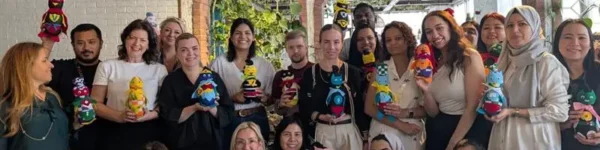 A group of people holding colorful, hand-crafted dolls, smiling together indoors with greenery in the background.