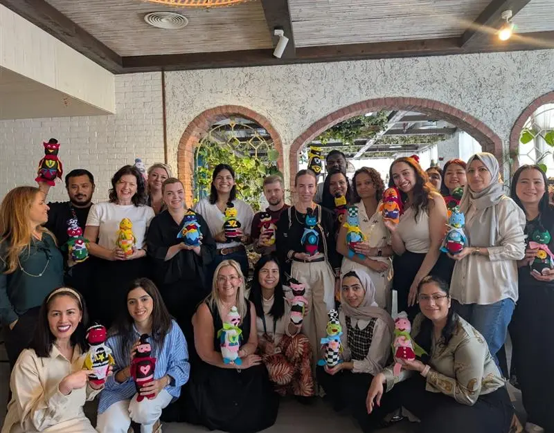 A group of people holding colorful, hand-crafted dolls, smiling together indoors with greenery in the background.