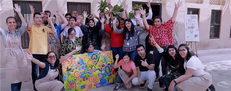 A diverse group of individuals smiles and celebrates joyfully, holding a colorful painted banner in a vibrant outdoor setting.
