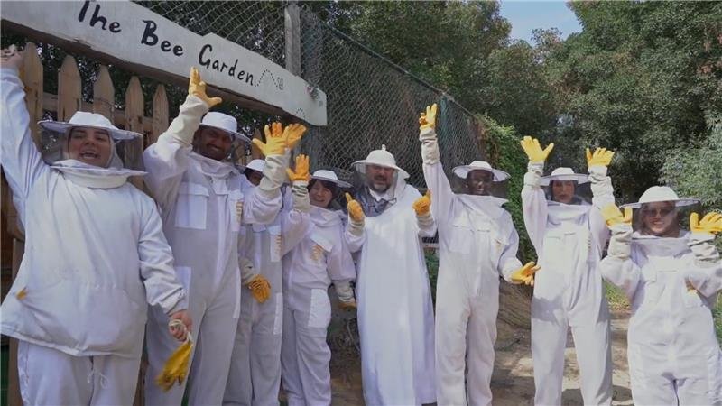 Group of people wearing beekeeping suits and yellow gloves, smiling and waving during an outdoor beekeeping activity