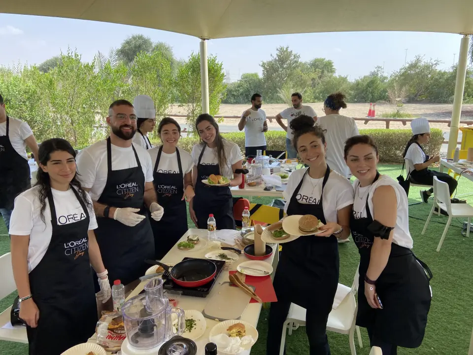 A group of smiling people wearing matching aprons and gloves stand in an outdoor setting. They are gathered around a table with cooking utensils and food, enjoying a team-building activity under a canopy, with greenery in the background.