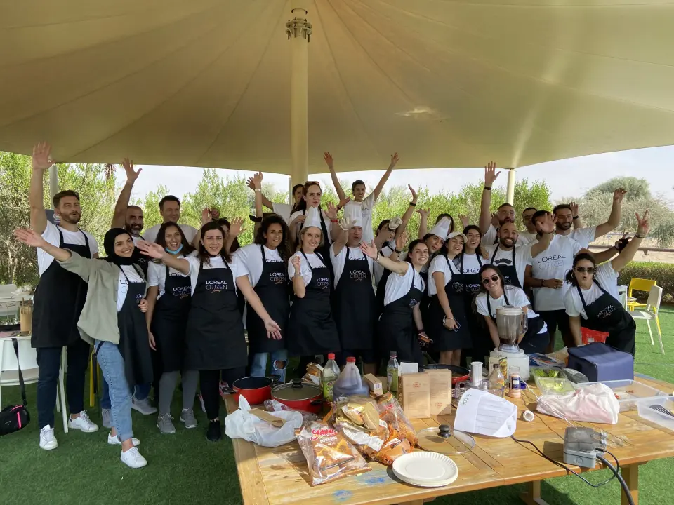 A group of people in matching aprons and shirts joyfully pose under a large canopy, with arms raised. A table with food and cooking items is in the foreground.